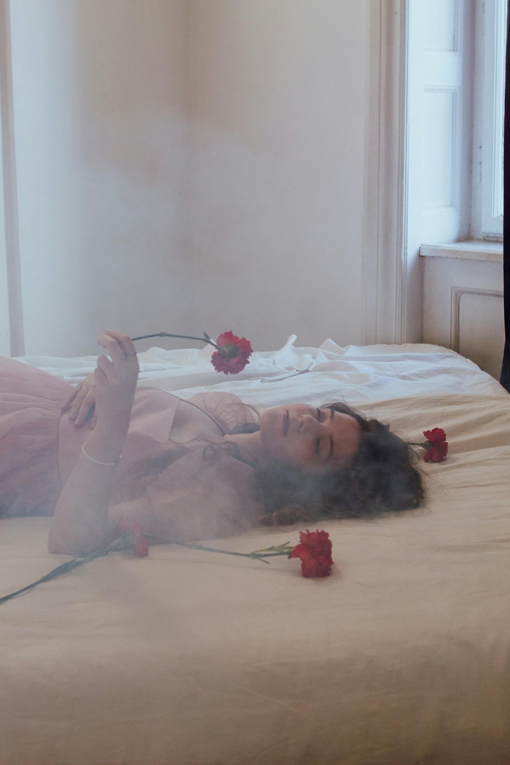 Young woman in a serene moment with red carnations on a bed, creating a moody and soft atmosphere.